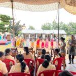 Colorful group of girls dancing in bright dresses on an outdoor stage under a canopy with an audience in beige uniforms watching from red chairs